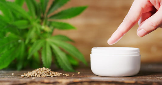 close-up of a female hand near a small tub of cream with a blurred hemp plant in the background.
