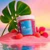 A jar of Bluebird Botanicals 'Lift' Tropical Punch Delta 9 THC Gummies sits on pink sand with a large green monstera leaf, a pink hibiscus flower, and red gummies, set against a tropical sunset background with water reflection.