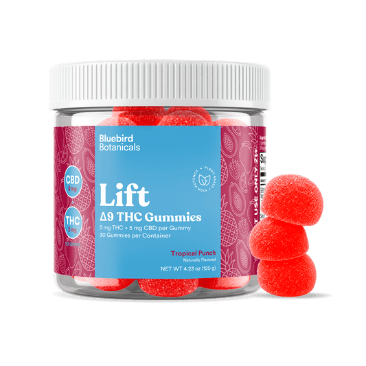 A clear plastic jar of Bluebird Botanicals Lift Tropical Punch Delta THC Gummies with a white ribbed lid and a blue and magenta label, shown alongside three red gummy candies on a white background.; Flavor: Tropical Punch;