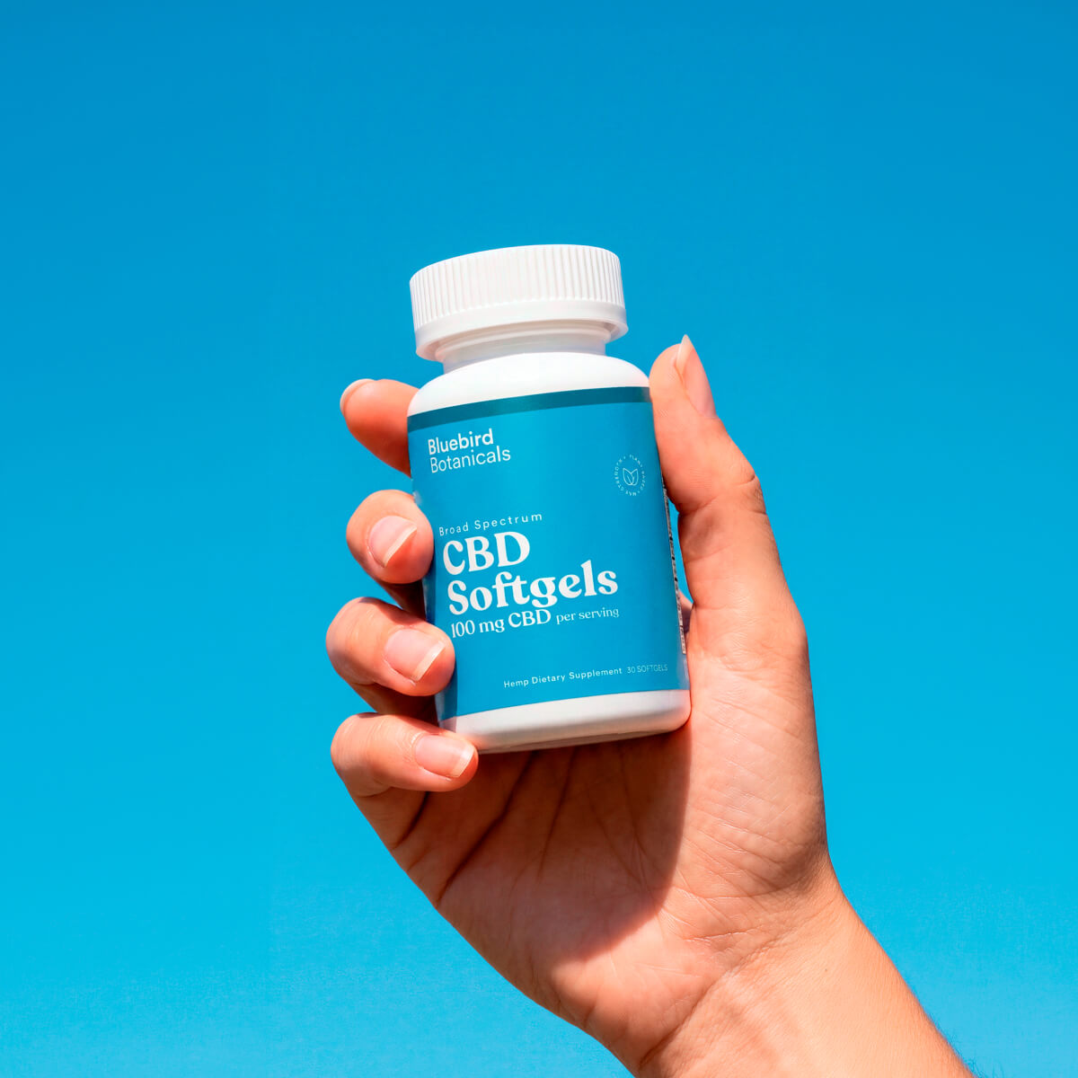 Hand holding a bottle of Bluebird Botanicals 100 mg Broad Spectrum CBD Softgels against a blue background.; Strength: 100 mg;