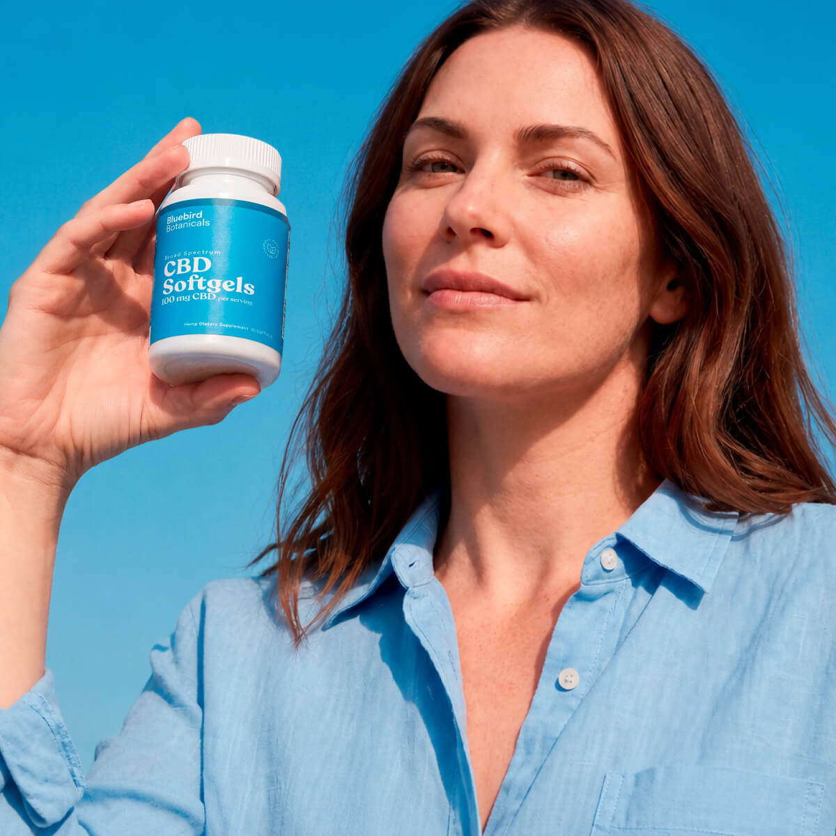Person in a blue shirt holding Bluebird Botanicals 100 mg Broad Spectrum CBD Softgels beside their face.; Strength: 100 mg;