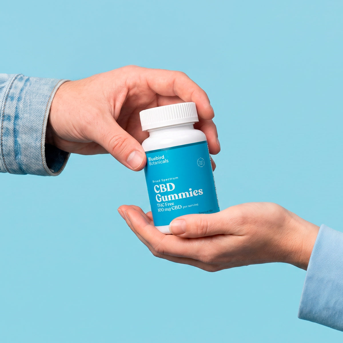 Two hands holding a bottle of Bluebird CBD Gummies on a blue background.; Strength: 100 mg;