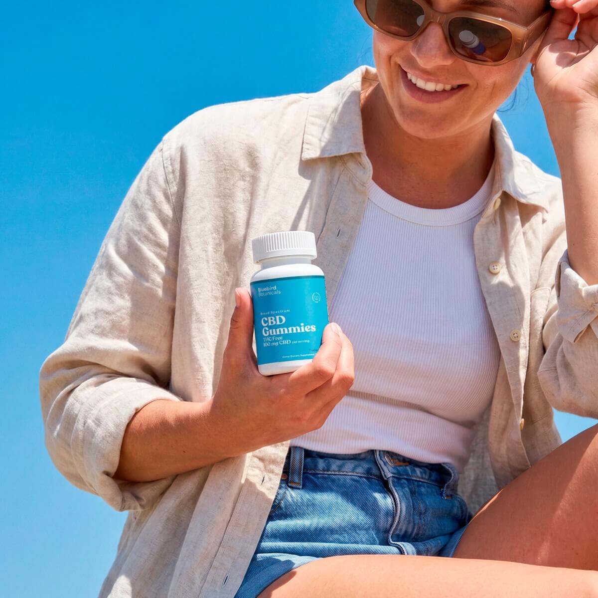 A woman outdoors under a bright blue sky holds a bottle of CBD Gummies. She is wearing a beige linen button-up shirt over a white ribbed tank top, denim shorts, and sunglasses.; Strength: 100 mg;