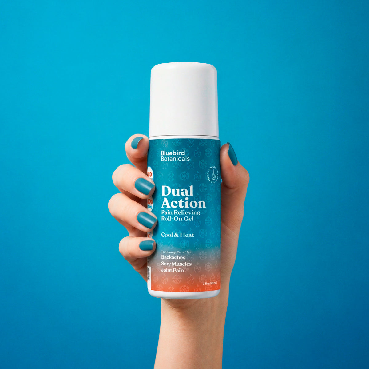 A hand with teal painted nails holds a roll-on bottle of Bluebird Botanicals Dual Action Pain Relieving Roll-On Gel against a solid bright blue background.
