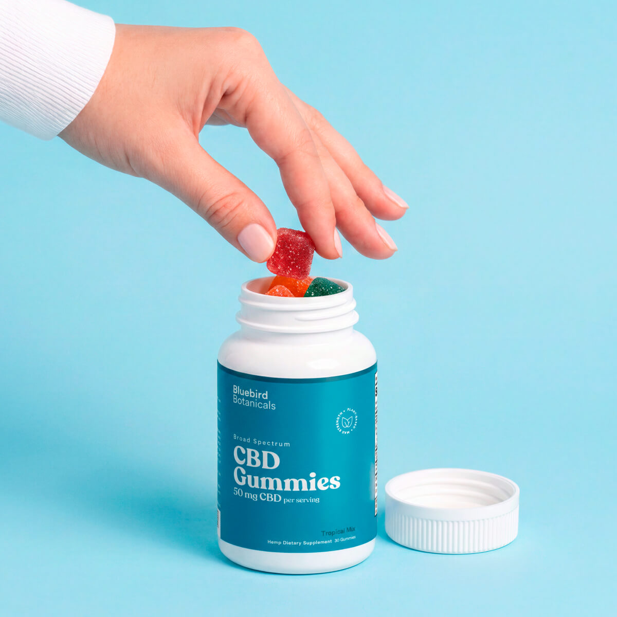 A hand wearing a white long-sleeve shirt is reaching into a white bottle of Bluebird Botanicals CBD Gummies, with one red gummy being lifted out. The bottle has a teal label and its white cap is placed beside it on a solid light blue background.; Strength: 50 mg;