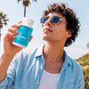 A man wearing sunglasses and a light blue button-up shirt holds up a white bottle of Bluebird Botanicals CBD gummies outdoors against a sunny sky with palm trees.; Strength: 50 mg;