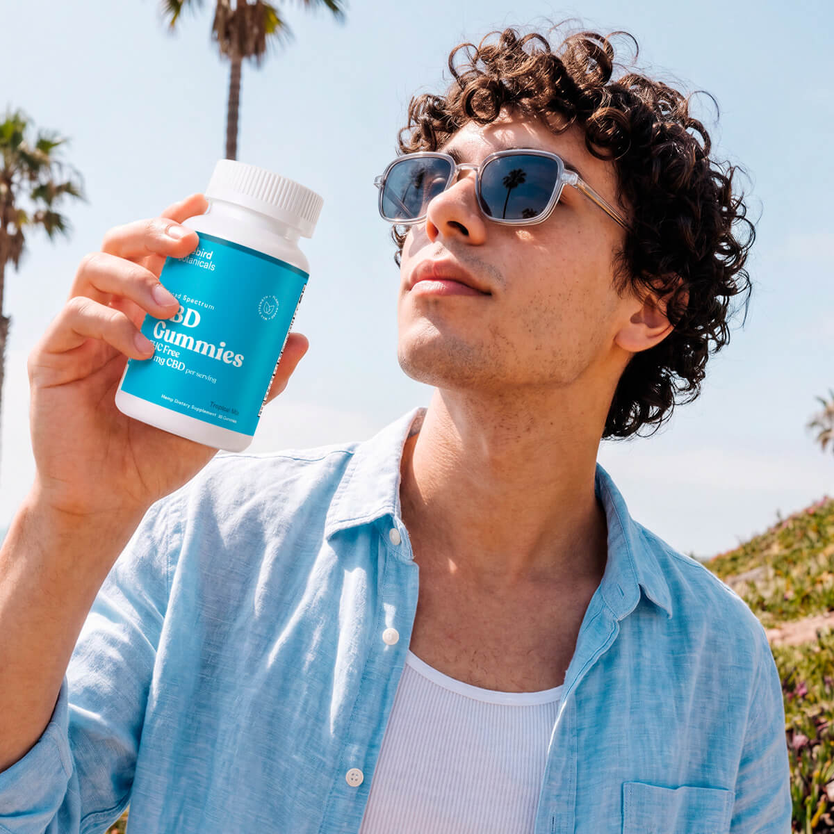 A man wearing sunglasses and a light blue button-up shirt holds up a white bottle of Bluebird Botanicals CBD gummies outdoors against a sunny sky with palm trees.; Strength: 50 mg;