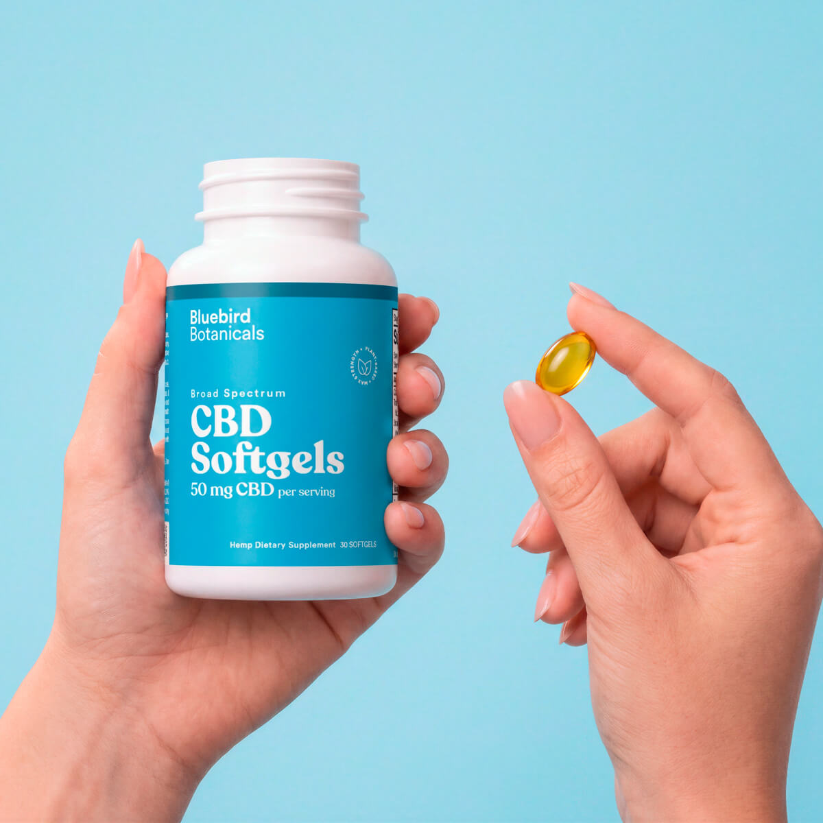 Hands holding an open bottle of Bluebird Botanicals 50 mg CBD Softgels and a single golden softgel.; Strength: 50 mg;