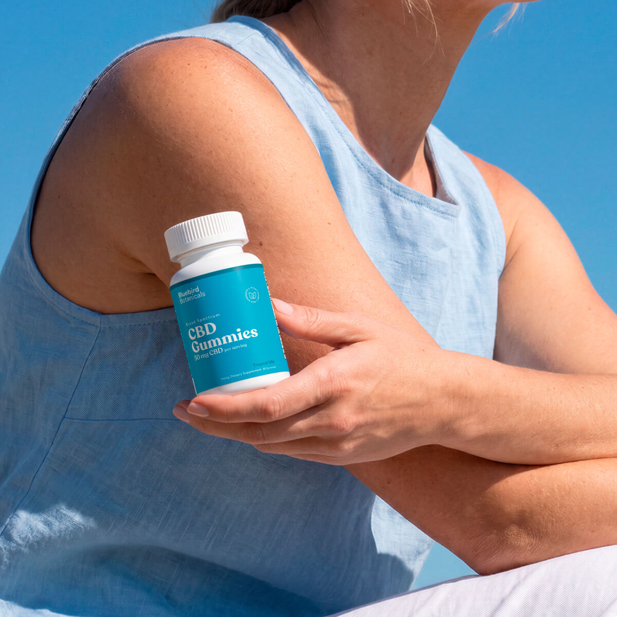 Person in a light blue sleeveless top holding a bottle of Bluebird Botanicals CBD Gummies.; Strength: 50 mg;